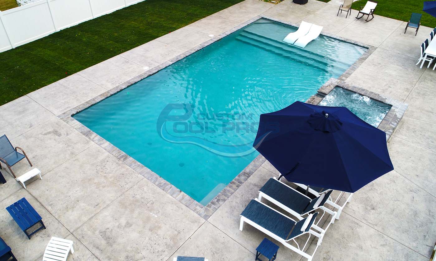 Aerial view of a modern swimming pool with clear blue water, surrounded by a stone deck, lounge chairs, and a large navy umbrella, set in a well-maintained backyard with green grass.