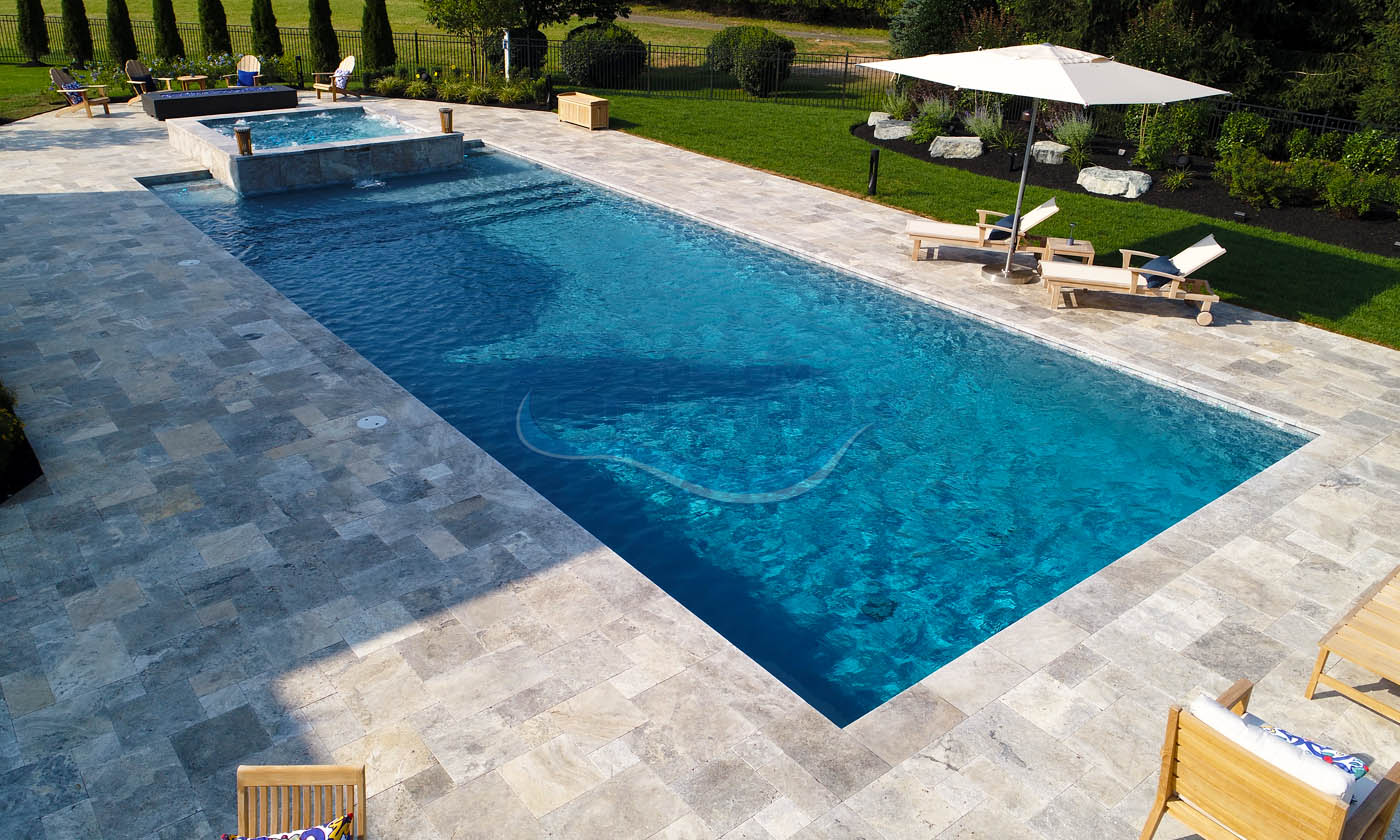 Aerial view of a luxurious swimming pool with crystal-clear water, surrounded by elegant stone paving and lush green landscaping, featuring sun loungers and a shaded area with an umbrella.