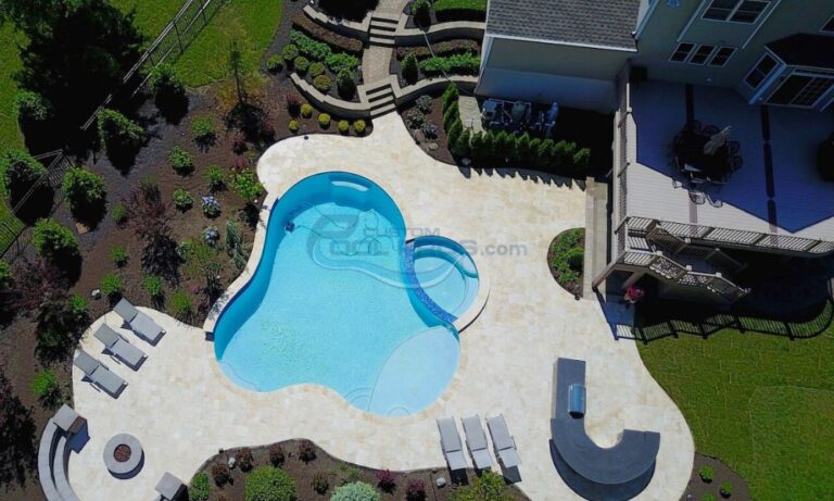 Aerial view of a beautifully landscaped backyard featuring a unique-shaped swimming pool, surrounded by stone patio, lounge chairs, and lush greenery, with a deck area visible on the right side, Swimming Pool Contractors NJ
