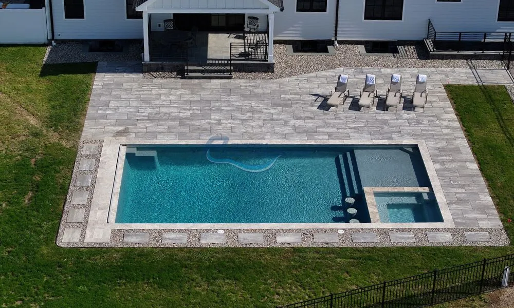 Aerial view of a modern backyard featuring a rectangular swimming pool surrounded by a spacious patio with gray stone tiles, lounge chairs, and a shaded seating area. The pool includes steps leading to a spa section, complemented by a well-maintained lawn, inground pools