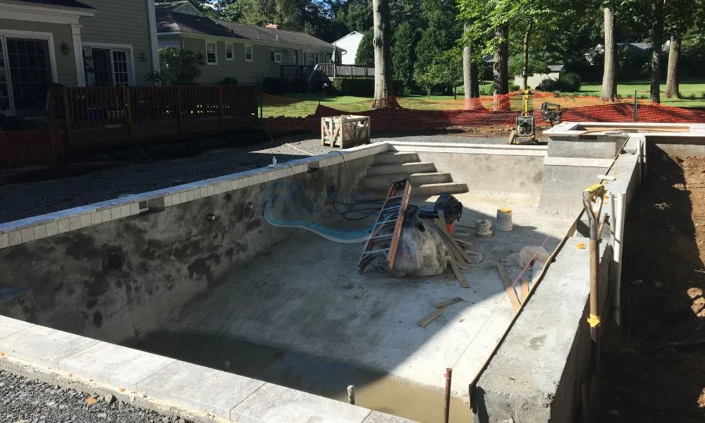 Construction site of an inground swimming pool, featuring a partially completed concrete structure with tools and materials scattered around. The background includes a house and trees, indicating a residential setting, custom swimming pools