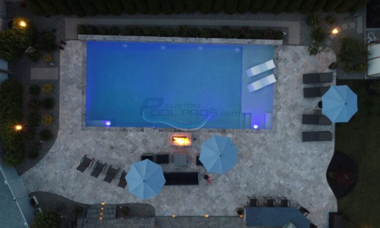 Aerial view of a modern swimming pool surrounded by stone decking, lounge chairs, and blue umbrellas, featuring ambient lighting and a fire pit, ideal for relaxation and outdoor gatherings, New Jersey Pool Companies
